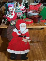 Tall hand-painted wooden Santa standing approximately 35 inches tall in front of a wooden table holding assorted Christmas-themed plates, a red decorative sleigh figurine, nutcracker figurines, glass bowl with red details, Hallmark teacups and service set, and various other holiday decor items.
