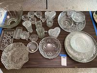 Wide view showing entire collection of clear glass items including punch bowl, cups, heart-shaped covered dishes, bowls, and platters arranged on a brown table.