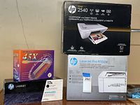 Photo of all four boxed items including HP Deskjet 2540 printer, HP LaserJet Pro M102w printer, HP 17A laser toner cartridge, Afreey 45X external CD-ROM drive boxed together on a table