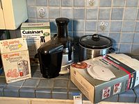 Overview photo showing Waring Automatic Juicer box, Cuisinart hand blender box and unit, Black & Decker waffle maker box, crockpot with lid, and Omega juicer unit on countertop.