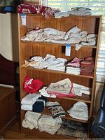Wooden shelving unit filled with folded linens including lace fabrics, sheets, red and white cloths, and some Christmas themed napkins and pot holders.