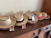 Front row of six straw hats on wooden surface, showing natural straw material and different colored ribbon bands, some with tags attached.