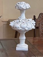 Full view of the tall white china fruit motif centerpiece on a wooden table, showing the two-tiered design and detailed fruit and leaf decorations.