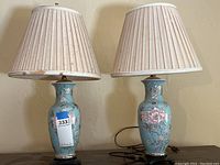 Two matching ceramic table lamps with pleated beige shades, both with floral patterns on light turquoise bases, placed side by side on a surface.