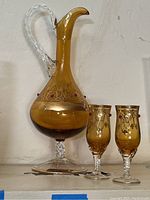 Full view of amber Venetian glass decanter with clear twisted handle, gold gilding, and red bead accents next to two matching glasses