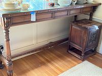 Full side view of the long tall vintage buffet table showing the carved wooden legs, rectangular glass top with some staining and decorative apron with inlaid panels and rosettes.