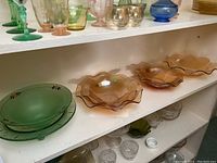 Shelf view of five carnival glass bowls including three amber iridescent scalloped/rippled edge bowls and two green glass bowls, one footed with etched starburst pattern and one shallow.