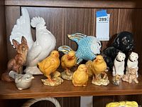 Group of ceramic animal figurines showing white rooster, chicks, fox, fish, dog and small cats, arranged on shelf.