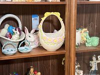 Shelf display of ceramic Easter decorations including a large white bunny figurine, a large scalloped Easter basket with yellow bow and daisies, smaller dishes, and other ceramic objects
