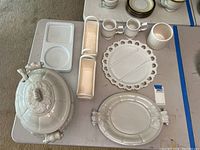 White ceramic covered soup tureen, platters, mugs, small trays, and planters arranged on table.