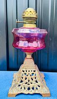 Front view of the antique cranberry glass oil lamp showing the ornate iron base, cranberry glass reservoir, and brass burner crown.