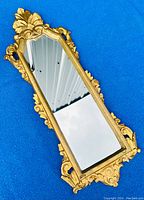 Photo showing the mirror laying flat on a blue surface with a gilded frame featuring leaf and scroll motifs and a narrow rectangular reflective glass panel.