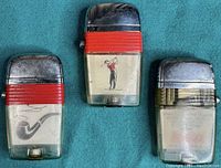 Three vintage Scripto-Vu lighters laid flat showing novelty inserts (golfer, pipe, machinery) with red ribbed band and chrome tops