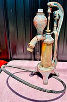 Photo showing old cast iron hand water pump standing on a purple surface with black background, revealing curved spout and circular base ring.