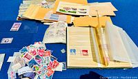 Wide view showing a large loose pile of various postage stamps, some still in packaging or albums, on blue background.
