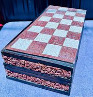 Closed view of foldable wooden chess board with red and white carved checker pattern and ornate carved edge detail.