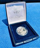 Opened clamshell case showing white satin inner lid with US seal and text '1999 SUSAN B. ANTHONY PROOF COIN' and the silver dollar proof coin inside plastic capsule on blue velvet lining.