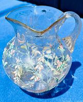 Clear blown glass pitcher with hand-painted enamel floral decoration of white and pink flowers with green stems and leaves, shown on a blue surface.