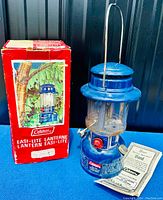 Front view of lantern standing next to its original red box and instruction pamphlets on blue cloth background.