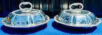 Pair of silver plated lidded serving dishes with scalloped edges and ornate removable handles viewed from an angle.