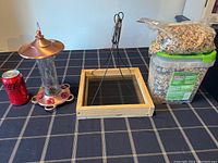 Full lot showing hummingbird feeder, seed tray bird feeder, plastic container and bag of bird seed, and reference soda can.