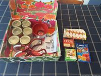 Photo of all canning supplies laid out in floral storage box on checkered tablecloth, showing jars, tools, pectin packets and labels.