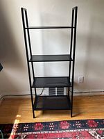 Front view of black metal bookshelf with four shelves, placed on wooden floor against white wall.