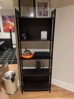 Tall dark wood IKEA Laiva bookcase with five shelves shown with miscellaneous objects on each shelf and a silver trash can beside it.