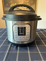 Front view of Instant Pot Digital Pressure Cooker showing control panel with cooking options and numeric display. Stainless steel body with black handles.