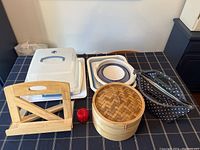Overview of the kitchen equipment on table including wood cookbook stand, bamboo steamer, cake carrier, large white tray, collapsible bowl and drying rack, and red kitchen timer.