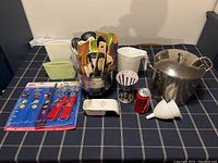 Photo showing the full assortment of kitchen utensils and accessories arranged on a table with a soda can for scale, including spatulas, ladle, tongs, vegetable peeler, scissors, measuring tools, storage bags, funnels, milk jug, and large stock pot.