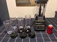 Ninja Auto iQ blender base with large blender jug attached and four individual smoothie cups with lids arranged on table. Coca-Cola can placed for size reference.