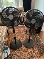 Two black Comfortmate standing floor fans side by side on a patterned rug indoors, showing full height and base.