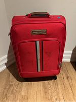 Front view of red Tommy Hilfiger carry-on suitcase showing brand patch, front zippered pocket, and vertical stripe design with minor stains.