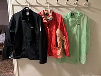 Three women's athletic jackets hanging on hangers, from left: black Helly Hansen, coral/peach Marmot, light green Nike