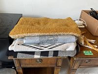 Stack of six scarves and shawls folded on a wooden nightstand, showing variety of patterns and colors including mustard yellow, gray, beige, black and white.