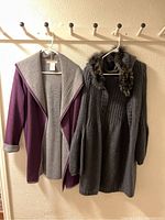 Two long women's sweater coats on hangers, one purple with gray shawl collar, the other dark gray with fur trim collar and buttons