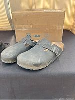 Pair of black Birkenstock slip-on sandals with visible wear and dust on the surface placed in front of a cardboard box.