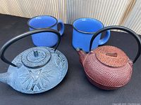 Pair of cast iron teapots and two blue enamel-look mugs on black surface.
