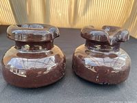 Two brown glazed telephone pole insulators shown side by side on a black surface reflecting light, highlighting their glossy finish and shape.