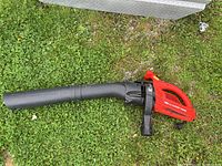 Full view of the leaf blower, showing red handle and black blower tube on grass.