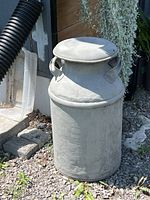 Full view of the vintage metal milk can outside next to a wall, showing overall shape, lid, and handles.