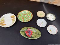 Overview of all ceramic items including the Moorcroft hibiscus ashtray, Trumpet plate, Ainsley plates, and assorted small decorative plates arranged on black surface.