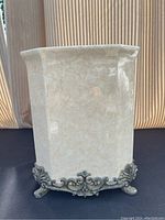 Front view of porcelain wastebasket showing marbled cream finish and ornate metal feet and trim.