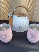 Wide shot showing white Rae Dunn tea kettle and two pink covered wine cups with lids, all labeled accordingly.