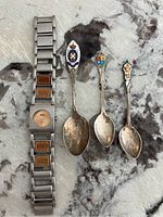 Photo showing three sterling silver souvenir spoons with enamel crests and a Ladies Roots wristwatch. The spoons have stamped markings and show tarnishing. The watch has a metal and leather blend band with 'Roots' logos.