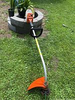 Full length view showing the trimmer laying on grass, curved shaft, orange cutting guard, and handle.