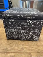 Front view of the square ottoman showing the black fabric cover with white French script pattern.