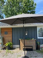 Full view of the tilting market umbrella in open position with black pole, rectangular shape and dark fabric canopy, set against a siding wall with plants.