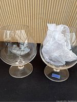 Two oversized clear Waterford Crystal cognac snifters photographed side by side on black surface with patterned background. One snifter contains tissue paper stuffing, the other empty showing wide bowl.
